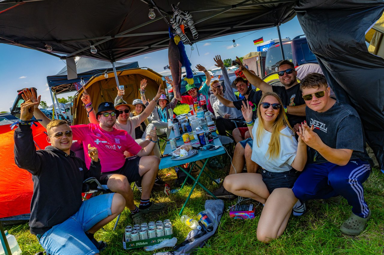 Eine Gruppe von Freunden sitzt unter einem Pavillon auf dem Campingplatz und streckt die Hände in die Luft.